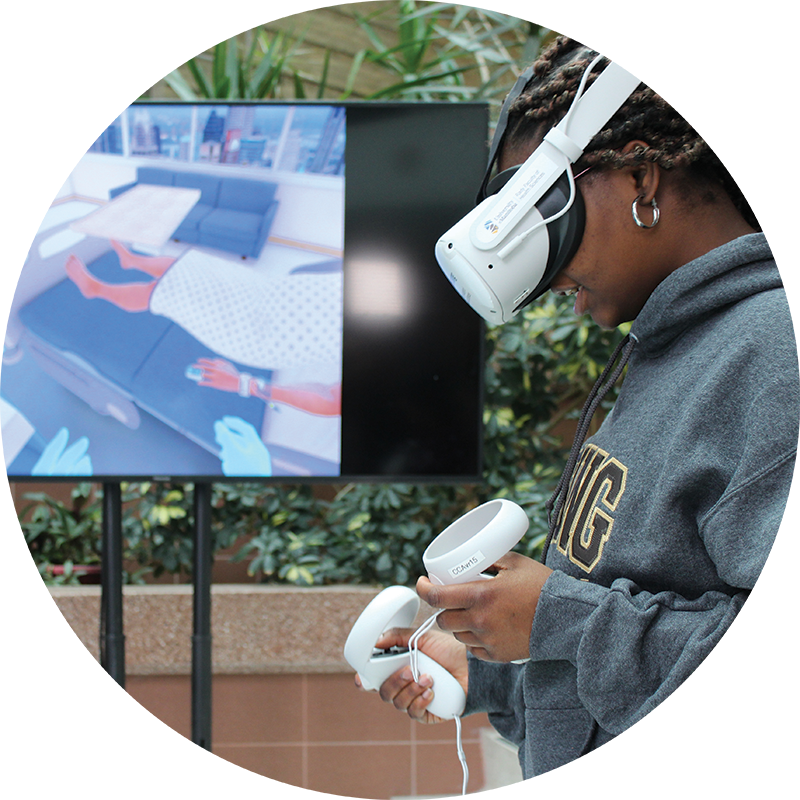 A person wearing a virtual reality headset and hand controls stands in front of a TV showing a simulated hospital environment.