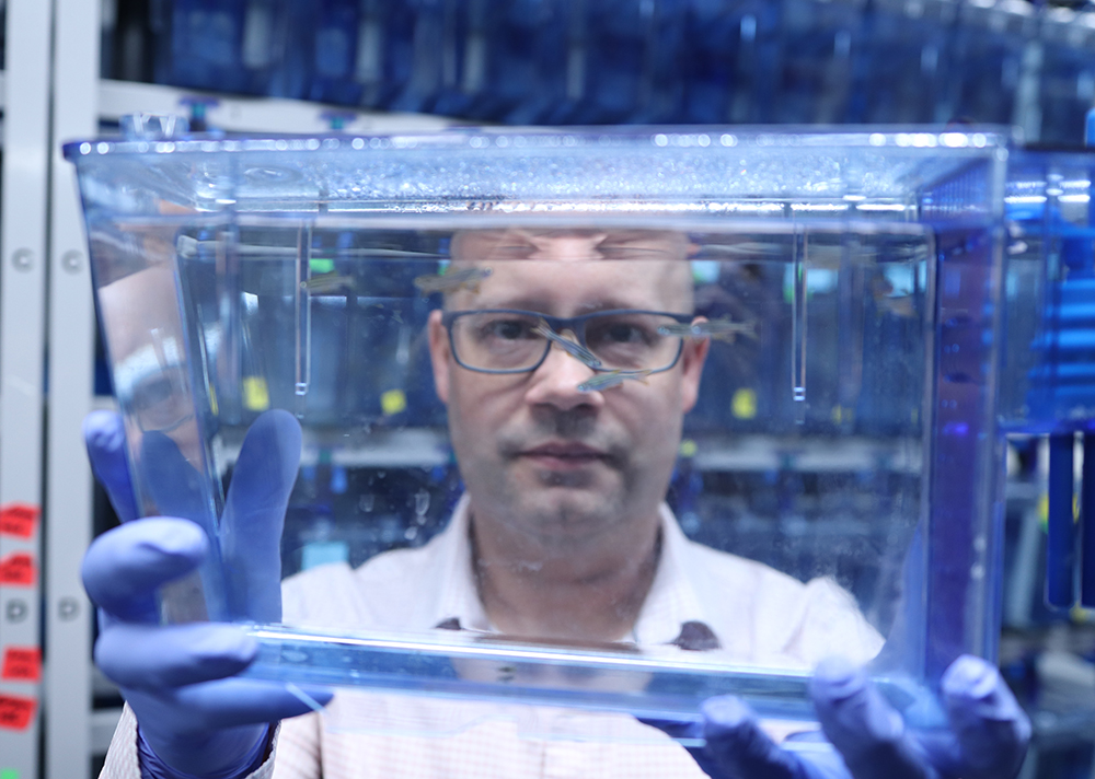 Dr. Benjamin Lindsey holds a small fish tank up to his face and looks through it. Fish are swimming in the tank.  