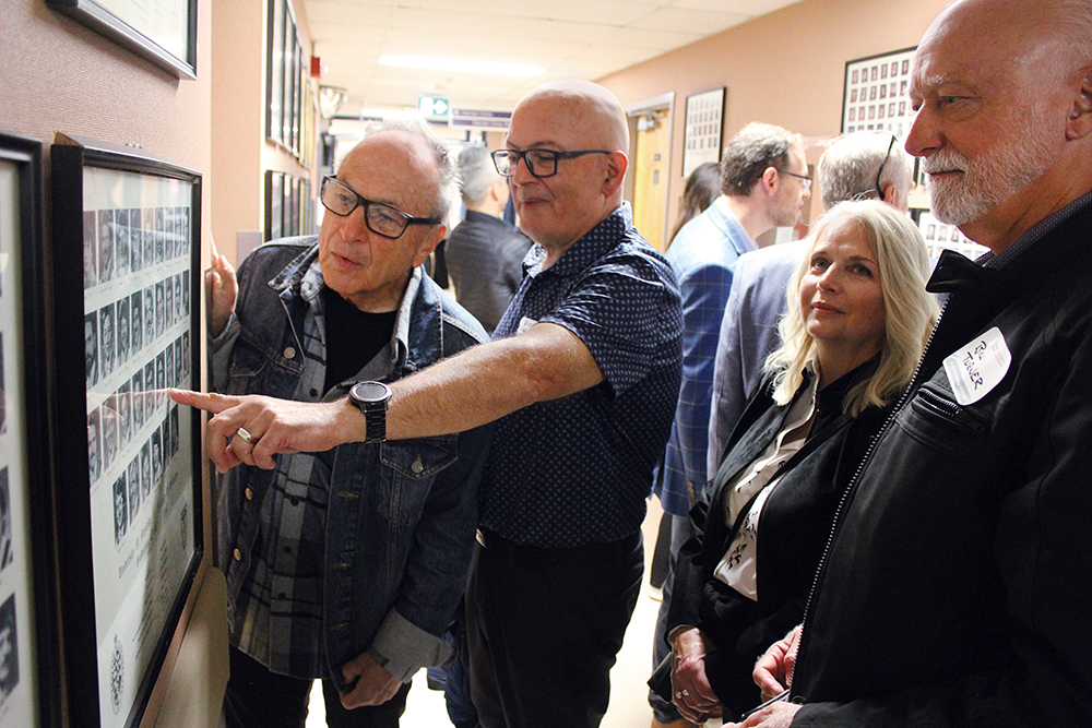 Four alumni look at framed graduation photos hanging on a wall. One alum is pointing at a photo. 