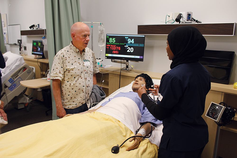 A student speaks with an alum. A patient care manikin lays on a bed in front of them.  