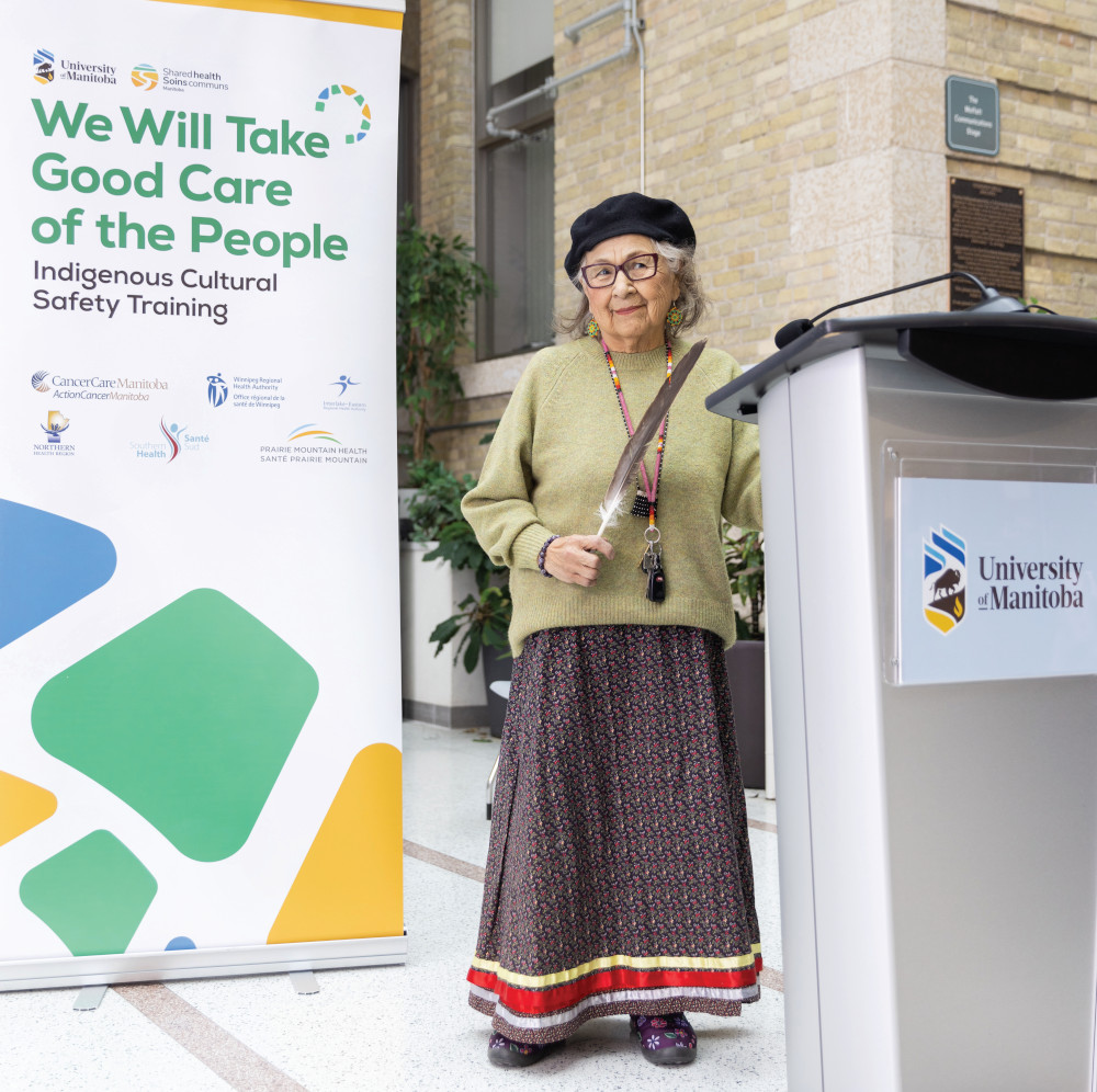 Elder Margaret Lavallee holds an eagle feather. She is standing behind a podium with the University of Manitoba log on the front. The main text on the banner behind her reads "We Will Take Good Care of the People. Indigenous Cultural Safety Training". 