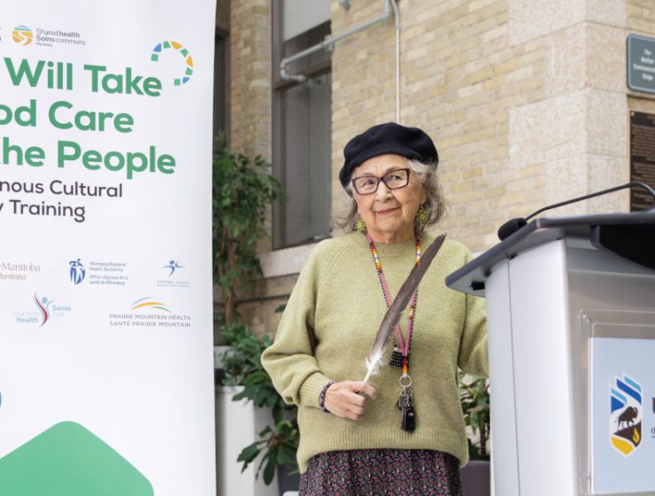 Elder Margaret Lavallee holds an eagle feather. She is standing behind a podium with the University of Manitoba log on the front. The main text on the banner behind her reads "We Will Take Good Care of the People. Indigenous Cultural Safety Training".