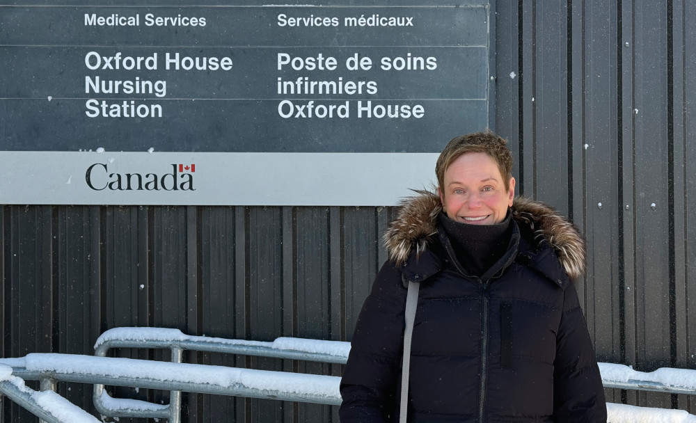 Dr. Judy Zetaruk stands outside wearing a parka. A sign behind her reads "Medical Services. Oxford House Nursing Station. Services medicaux. Poste de soins infirmiers Oxford House. Canada." 