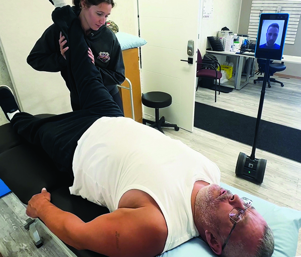 A rehab assistant works with a client while a physiotherapist watches on through a mobile tablet.