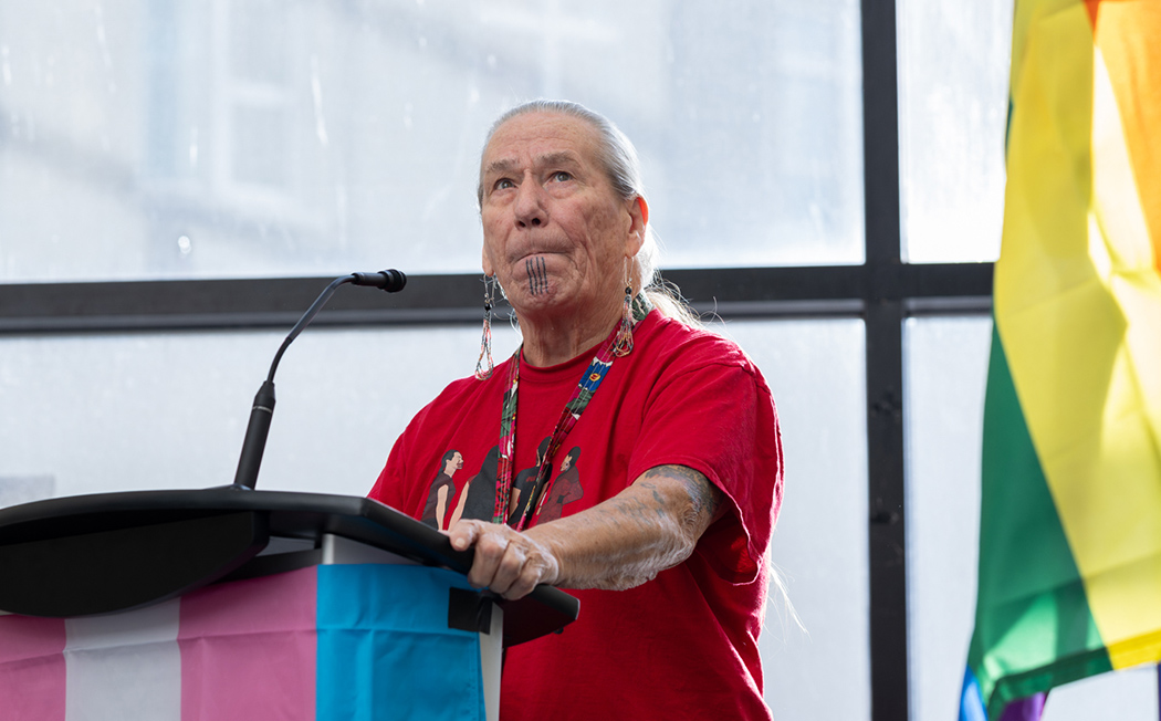 Elder Charlotte Nolin speaks in front of a podium that is draped in a transgender flag.