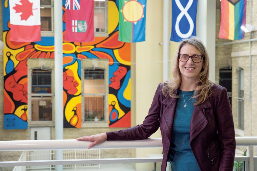 Dr. Sharon Bruce smiling at the camera, in the background are flags of Canada, Manitoba, Treaty 1, Métis and 2SLGBTQIA+.