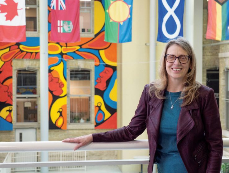 Dr. Sharon Bruce smiling at the camera, in the background are flags of Canada, Manitoba, Treaty 1, Métis and 2SLGBTQIA+.