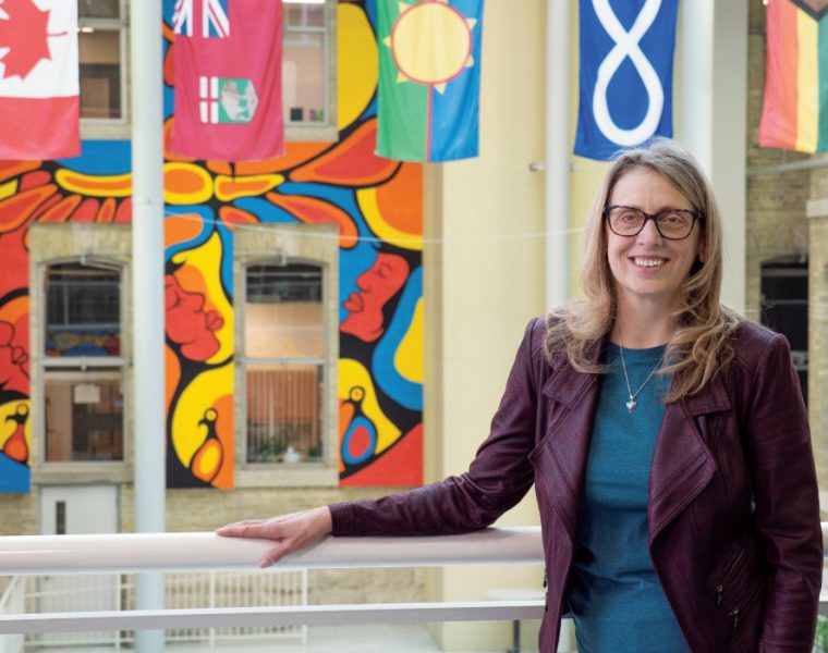 Dr. Sharon Bruce smiling at the camera, in the background are flags of Canada, Manitoba, Treaty 1, Métis and 2SLGBTQIA+.