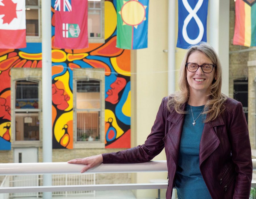 Dr. Sharon Bruce smiling at the camera, in the background are flags of Canada, Manitoba, Treaty 1, Métis and 2SLGBTQIA+.