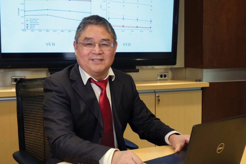 Dr. Depeng Jiang smiles at the camera behind and open laptop. Behind him is a TV screen.