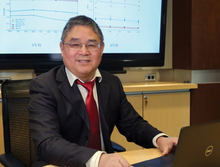 Dr. Depeng Jiang smiles at the camera behind and open laptop. Behind him is a TV screen.