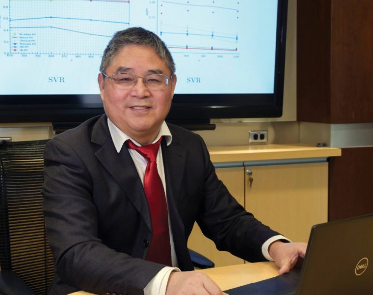 Dr. Depeng Jiang smiles at the camera behind and open laptop. Behind him is a TV screen.