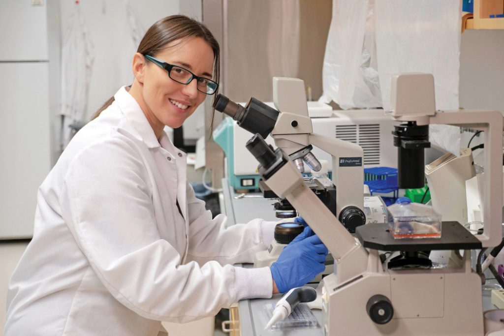 A scientist in front of a microscope in a lab, smiling at the camera.