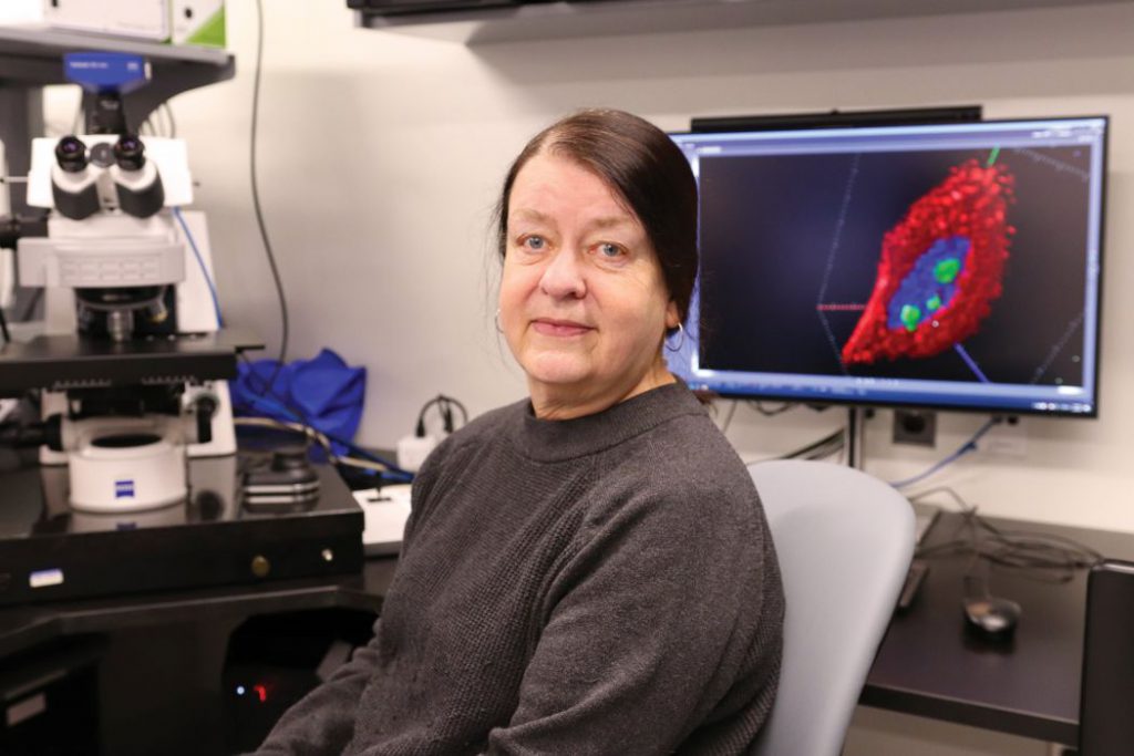 Dr. Sabine Mai sit in the imagine facility. A microscope and a computer screen with a graphic on it is behind her.