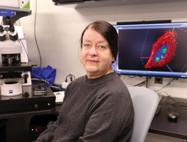 Dr. Sabine Mai sits in the imagine facility. A microscope and a computer screen with a graphic on it is behind her.
